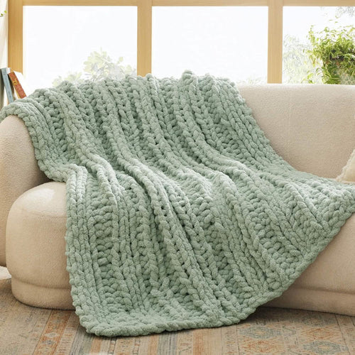 Sage green chunky knit chenille blanket with braided texture draped over a cream sofa.