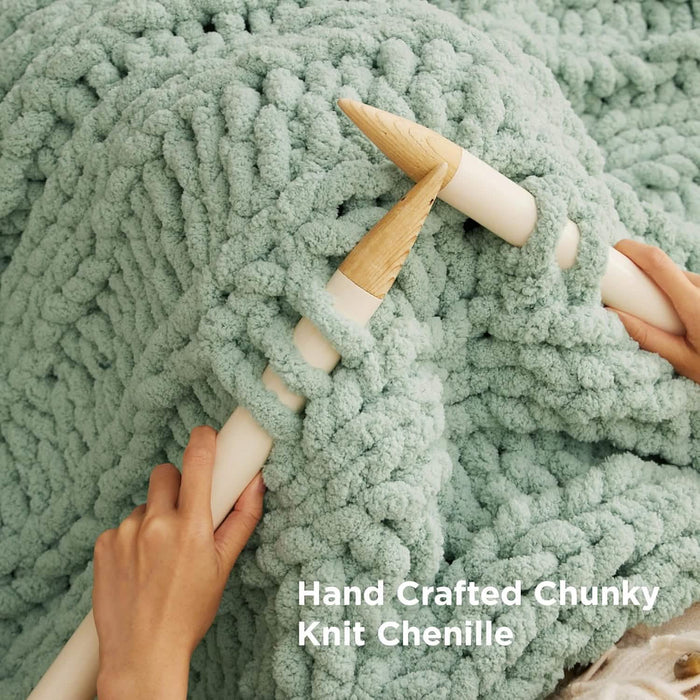 Sage green chunky knit chenille blanket being hand-crafted with large wooden needles, emphasizing plush texture and quality.