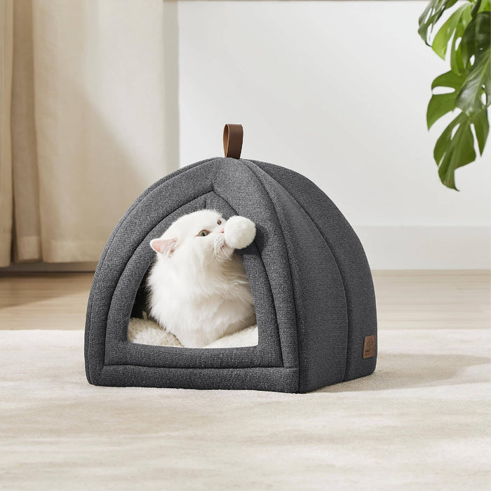 Dark gray faux-linen cat tent, fluffy white cat plays with pom-pom toy inside. Plush interior, faux-leather handle.