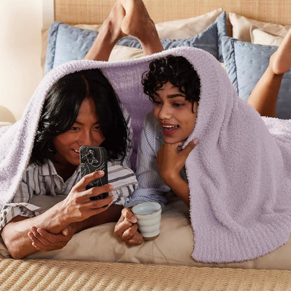 Two people sharing light lavender GentleSoft feather yarn blanket on bed, one with phone, other with mug.