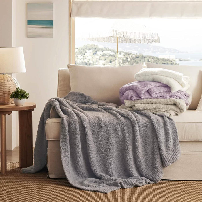 GentleSoft feather yarn throw blanket light gray draped on cream sofa, with stacked lilac, white, and gray blankets.