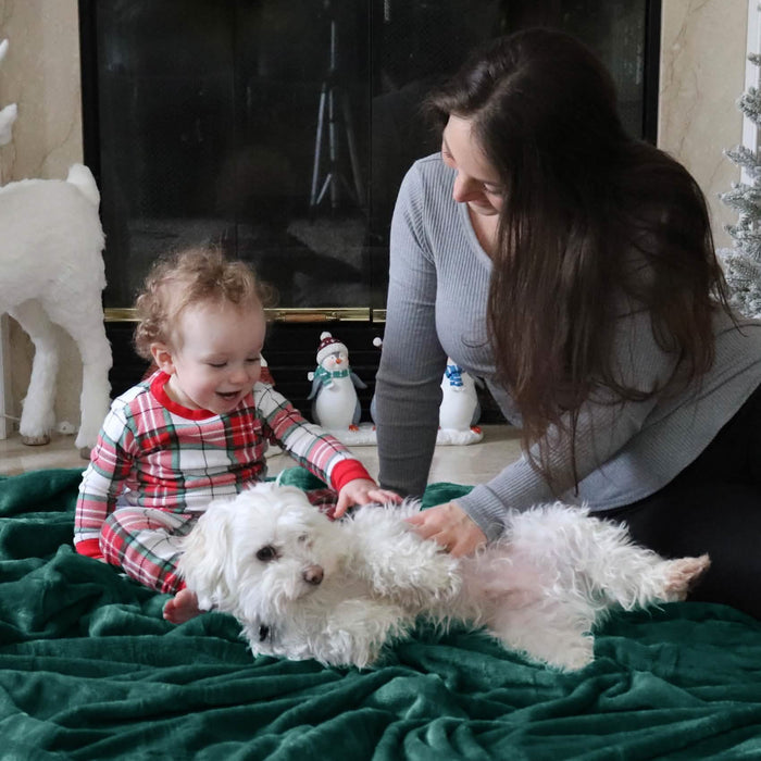 Woman and toddler in plaid pajamas petting white dog on dark green GentleSoft flannel blanket by fireplace.