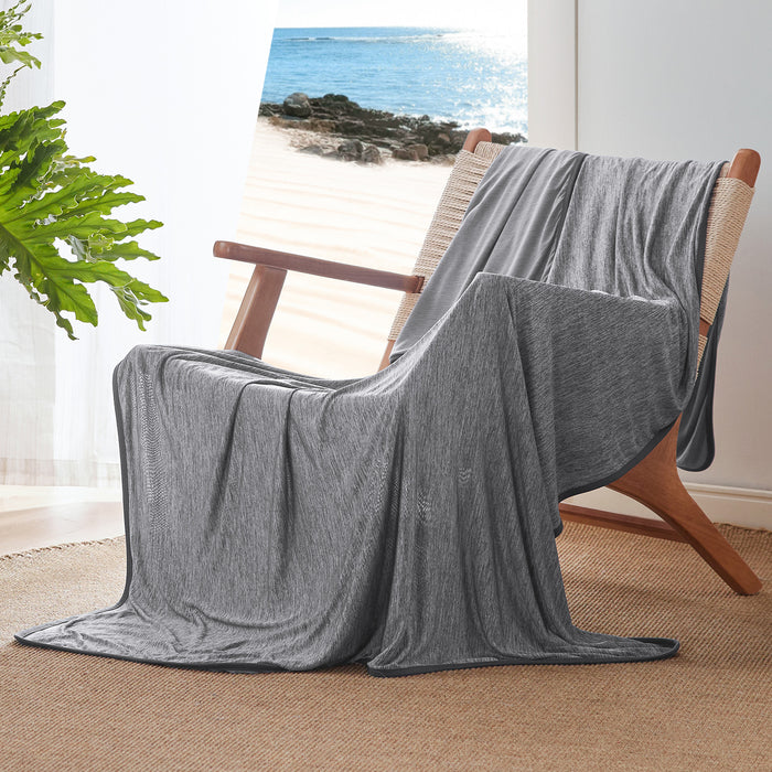 Gray GentleSoft reversible cooling blanket, nylon material, draped on a chair with a serene beach view.